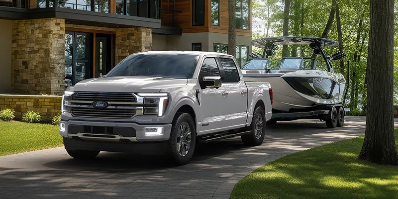 Light gray Ford F-150 truck towing a boat on a driveway, with a modern stone house and trees in the background.
