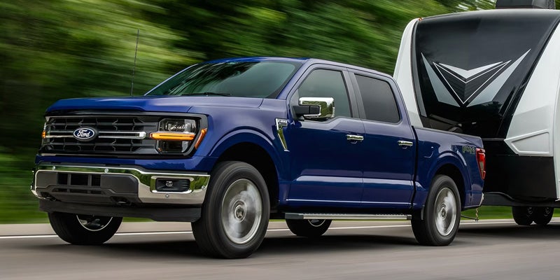 Blue Ford F-150 pickup truck towing a white and black travel trailer on a road, with motion blur.