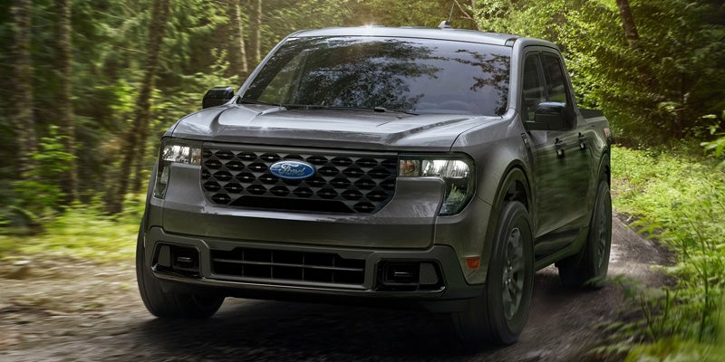 Dark gray Ford Maverick pickup truck driving on a dirt road through a green forest, blurred for motion.