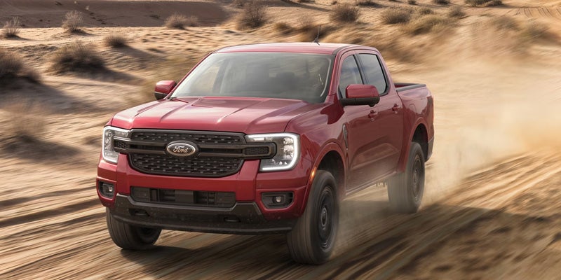 Red pickup truck driving through sand dunes, kicking up dust.